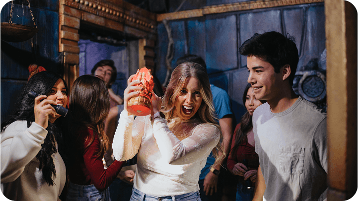 Group of friends celebrating indoors; a woman in white holds a trophy and shouts joyfully, while others smile around her.