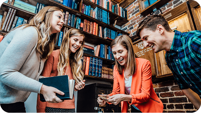 Four young adults gather around a table, smiling and examining a card. Behind them is a library-like setting with bookshelves.